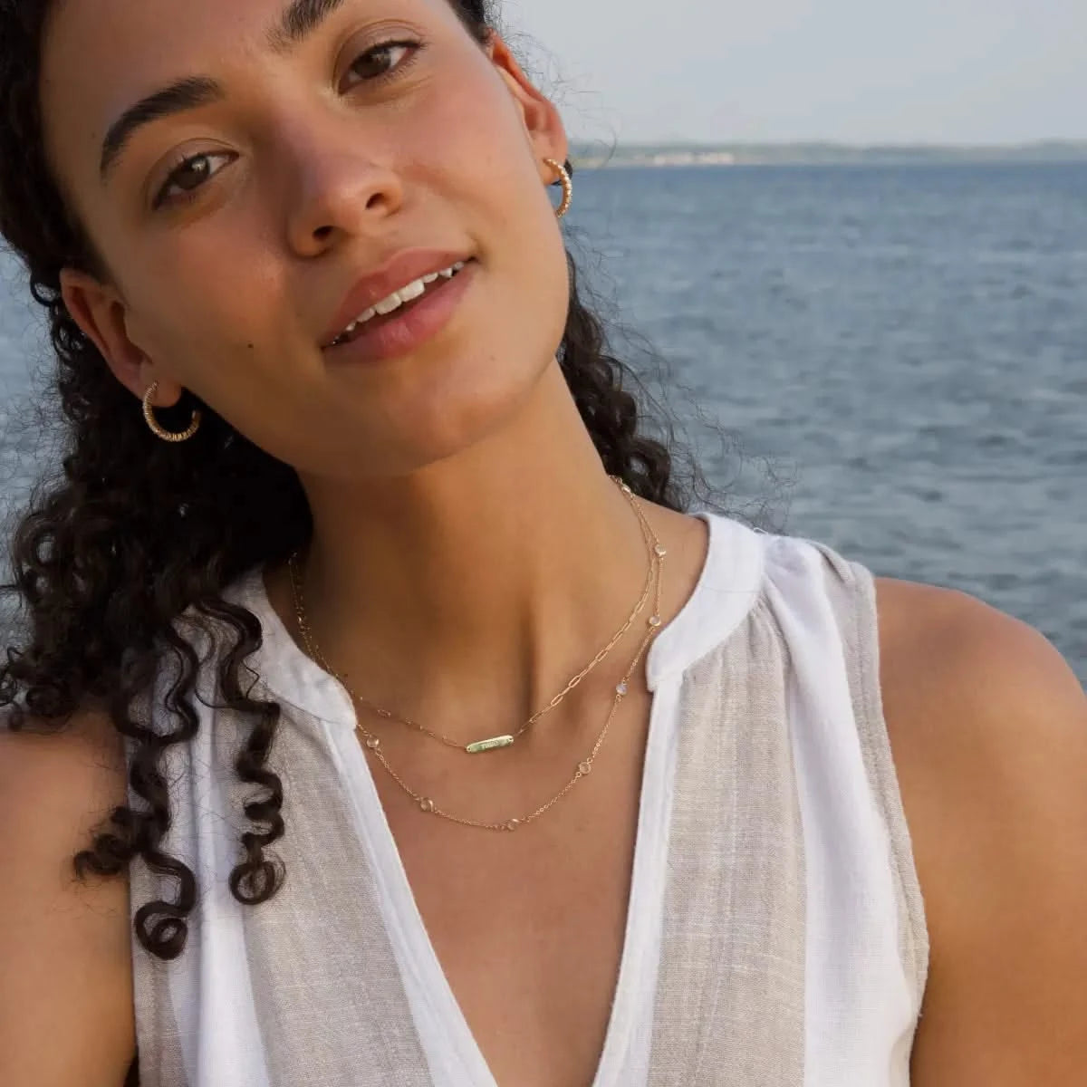 Woman wearing 1 Nameplate Necklace on Adelaide Mini paperclip chain, Bayberry Moonstone Necklace, and Rosecliff Birthstone Hoop Earrings, all in 14k yellow gold, styled near the ocean.