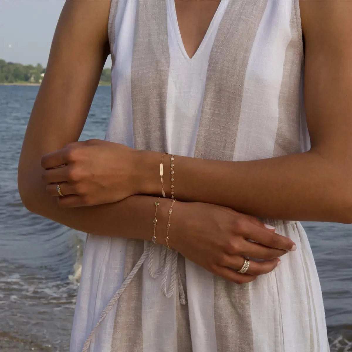 Woman near the ocean showcasing 14k yellow gold 3 Nameplate, Newport Opal, Bayberry Moonstone, and Grand Birthstone bracelets.