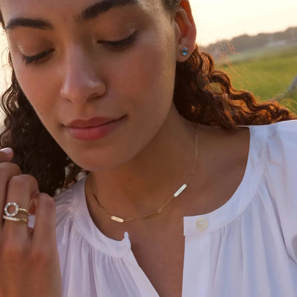 Woman standing by coastal greenery wearing a 14k gold Adelaide Mini chain necklace with four flat engraved nameplates, each personalized with her loved one