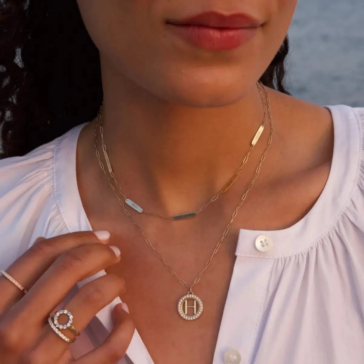Woman wearing 5 Petite Nameplate Necklace and Letter & Diamond Pendant in 14k gold, styled together near the ocean.