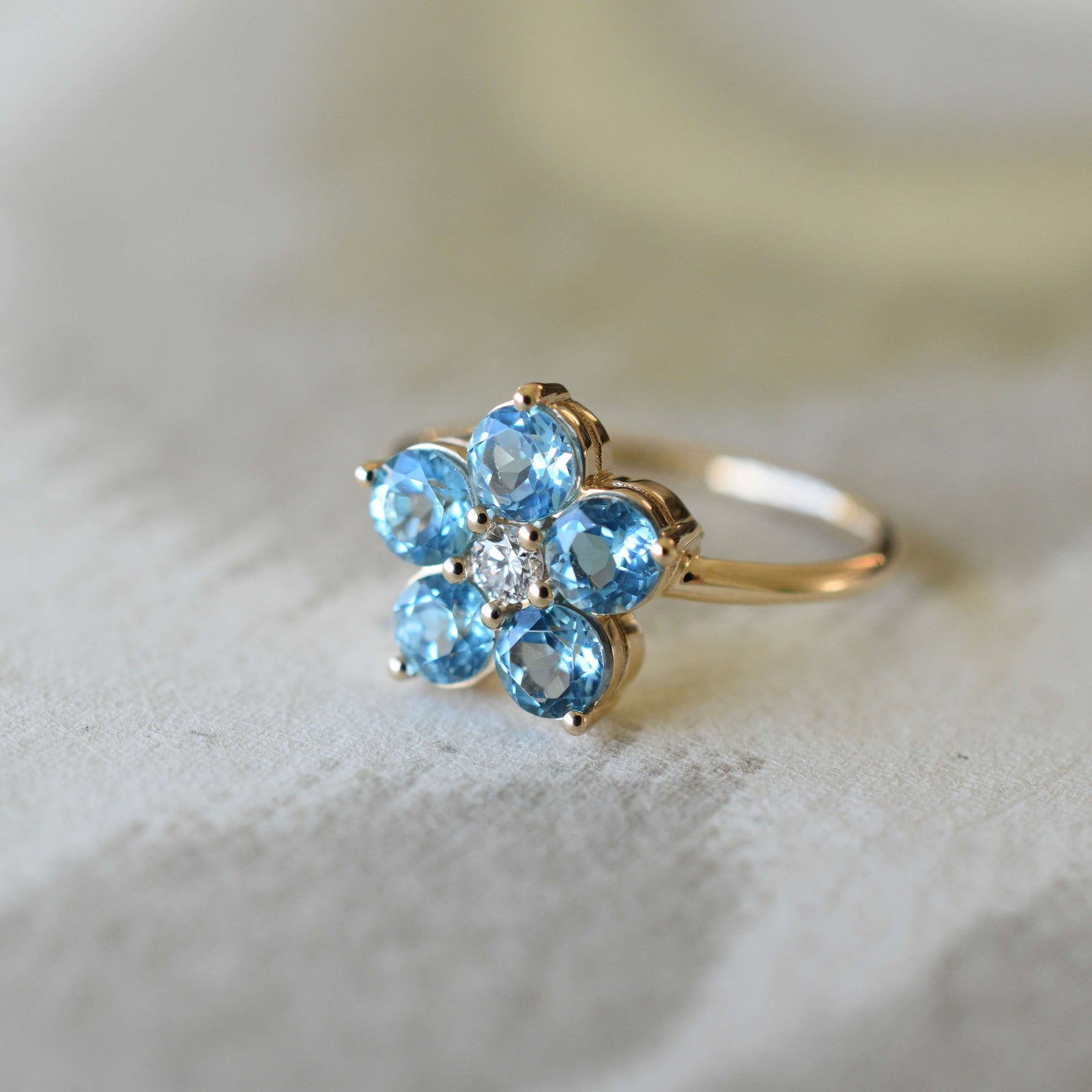 The Greenwich Nantucket Blue Topaz and Diamond Petal Ring features five prong-set blue topaz stones in 14k gold around a central diamond.