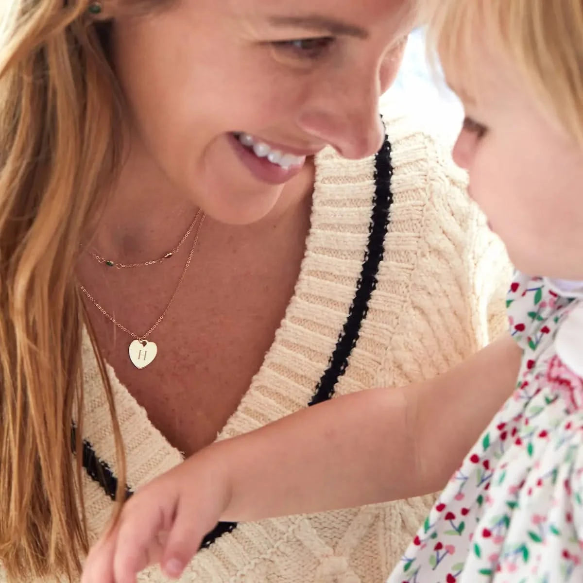 Woman smiles at her child while wearing a personalized gold heart initial necklace layered with a multistone chain.