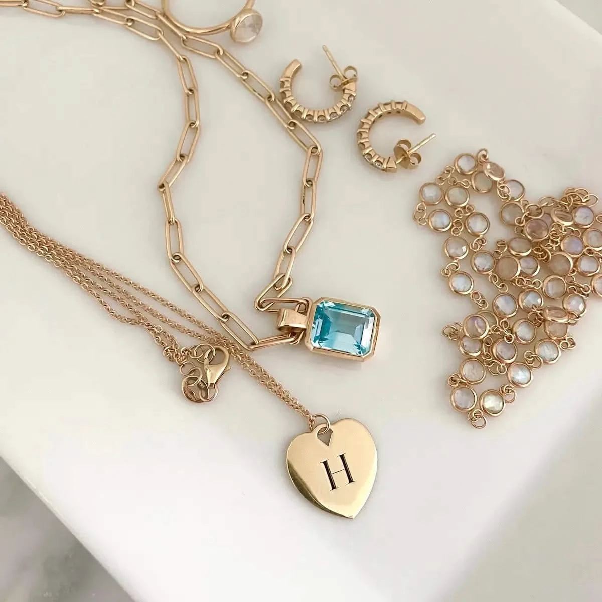 Flat lay of gold jewelry featuring a heart pendant engraved with "H", a blue gemstone necklace, and gold hoop earrings.