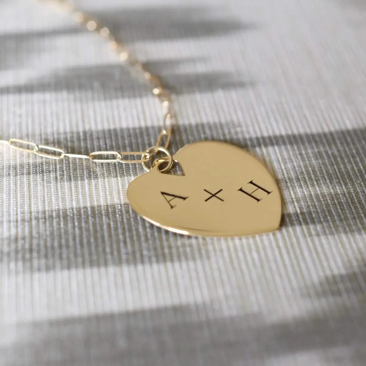 Close-up of gold heart pendant engraved with “A + H”, resting on striped fabric.