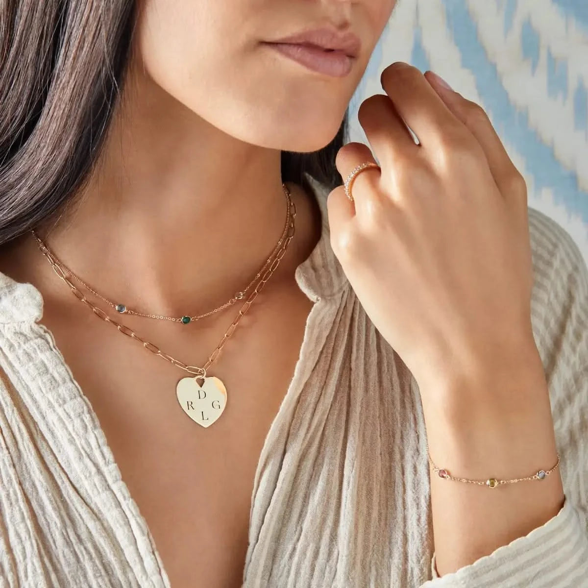 Woman wearing layered yellow gold heart pendant engraved with “D R G L” and a birthstone bracelet.