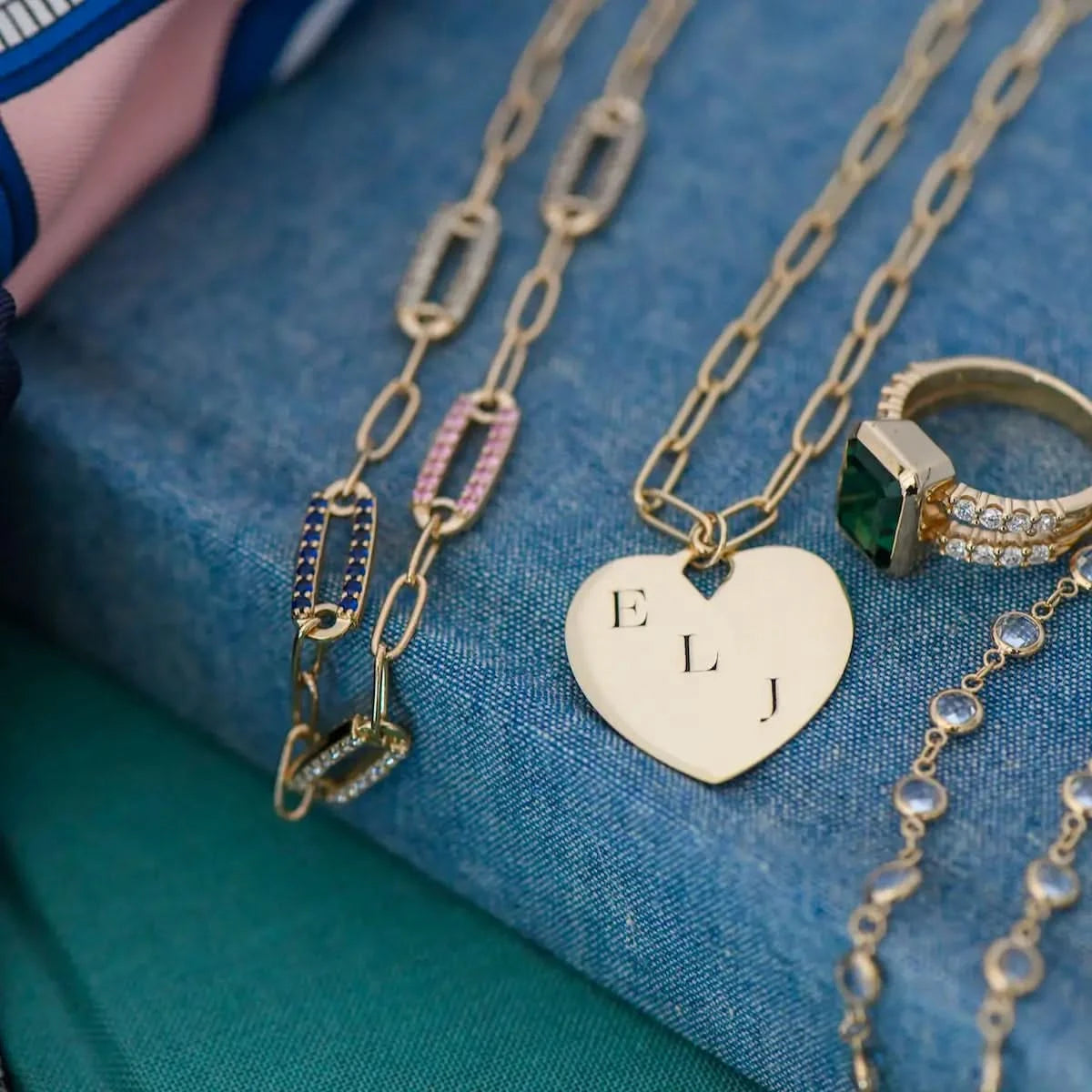 Engravable Large Flat Heart Pendant in 14k gold with initials E, L, and J, styled with Haverhill bracelets and rings on a denim-textured surface.