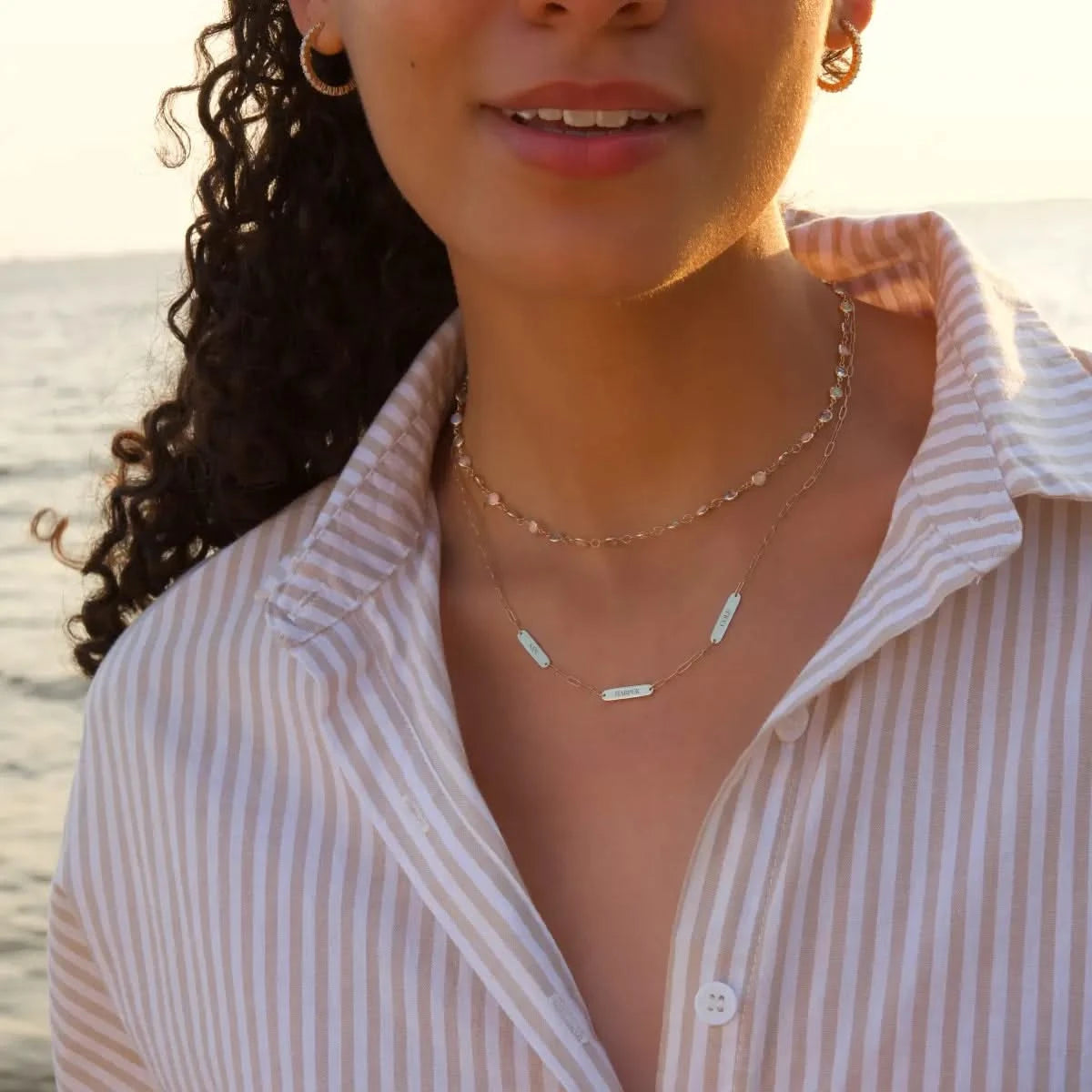 Woman standing by the ocean wearing 3 Petite Nameplate Necklace on Adelaide Mini paperclip chain in 14k gold, personalized with three engraved nameplates, layered with the Newport Opal Necklace in 14k gold, showcasing delicate opal gemstones.