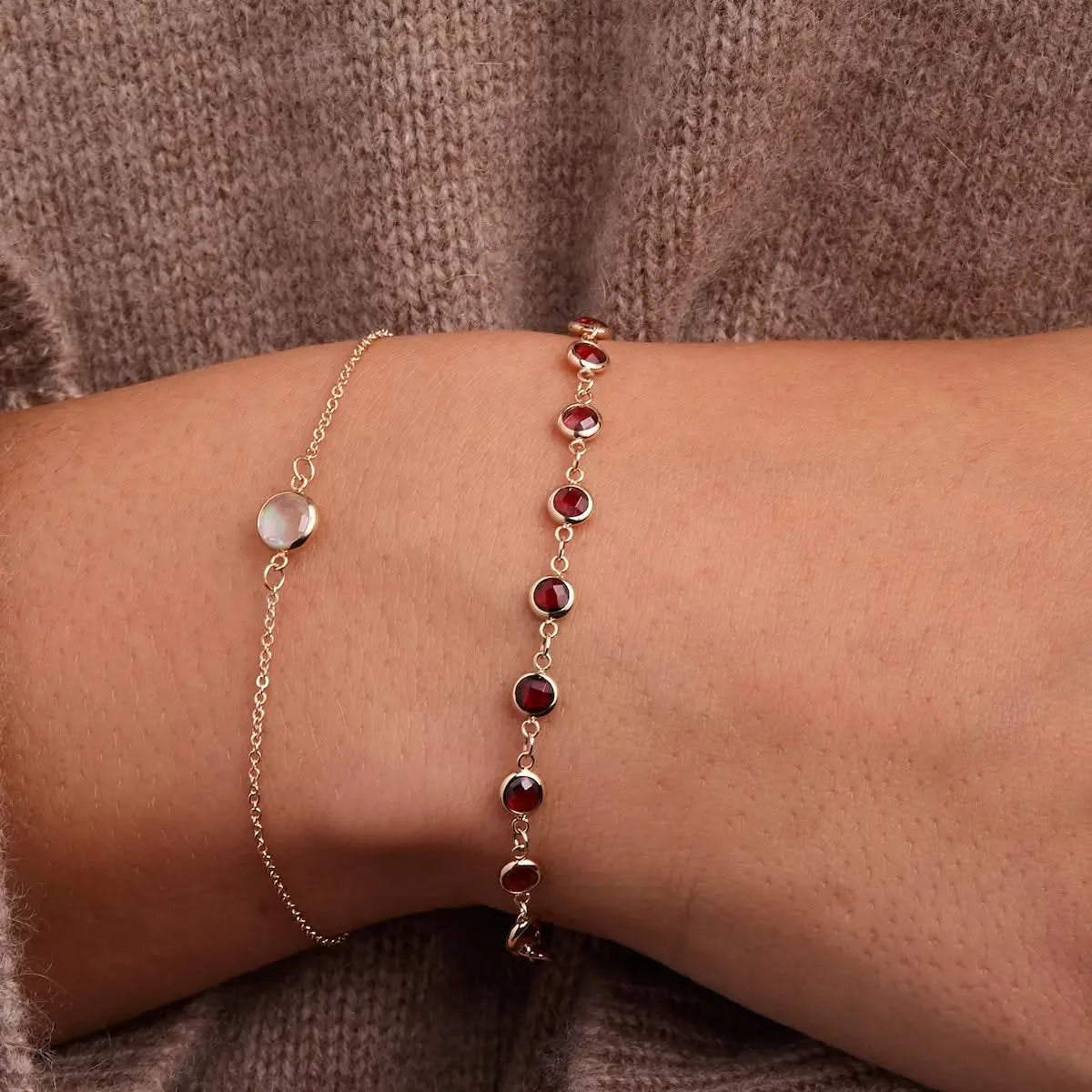 Close-up of woman wearing layered 14k gold birthstone bracelets, featuring a garnet bezel-set station bracelet and a minimalist moonstone chain bracelet.