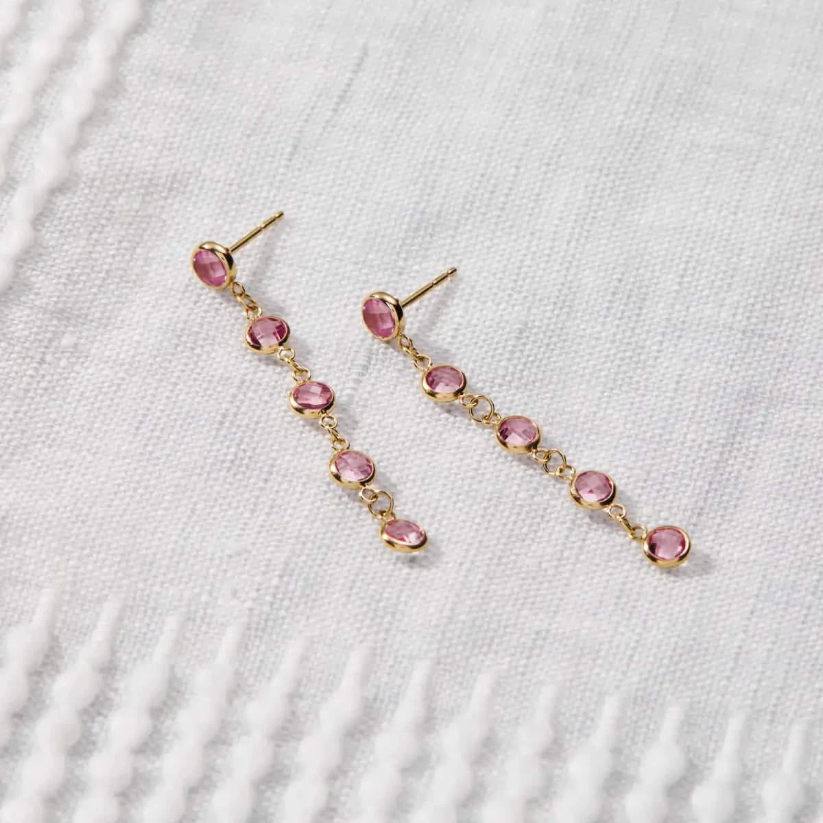 Newport Pink Sapphire Earrings in 14k gold are displayed on fabric, featuring five bezel-set briolette-cut Pink Sapphires.