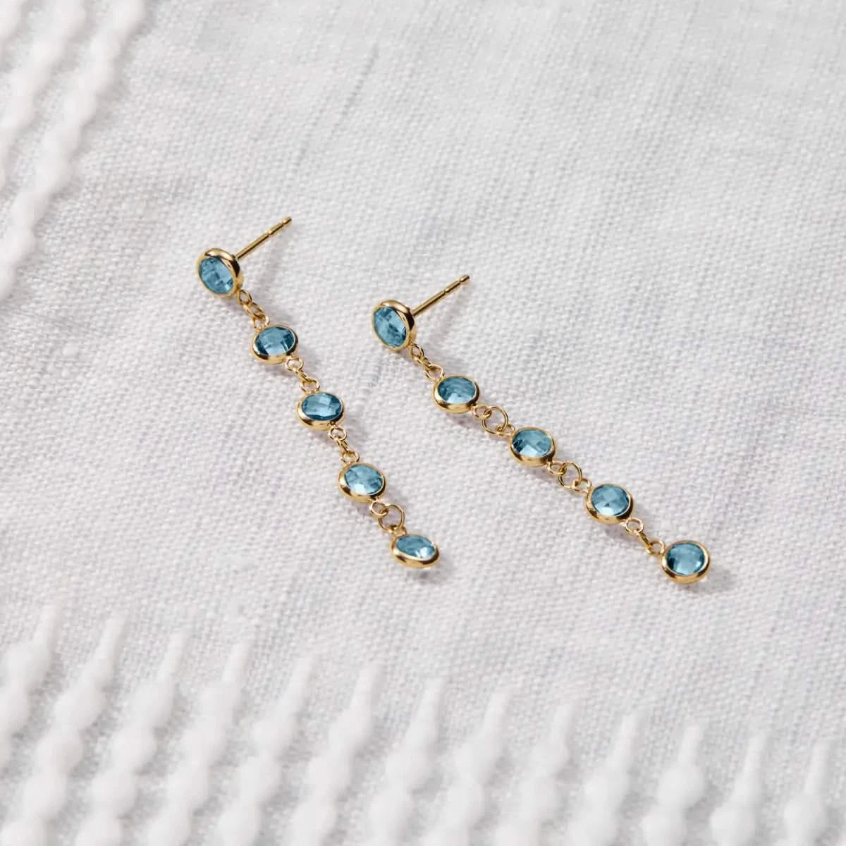 Newport Nantucket Blue Topaz Earrings in 14k gold are displayed on fabric, featuring five bezel-set briolette-cut Blue Topaz birthstones.