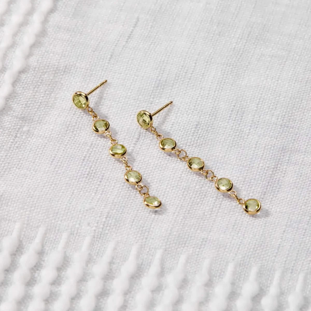 Newport Peridot Earrings in 14k gold are laid flat to highlight the five bezel-set August birthstones in a linear drop style.