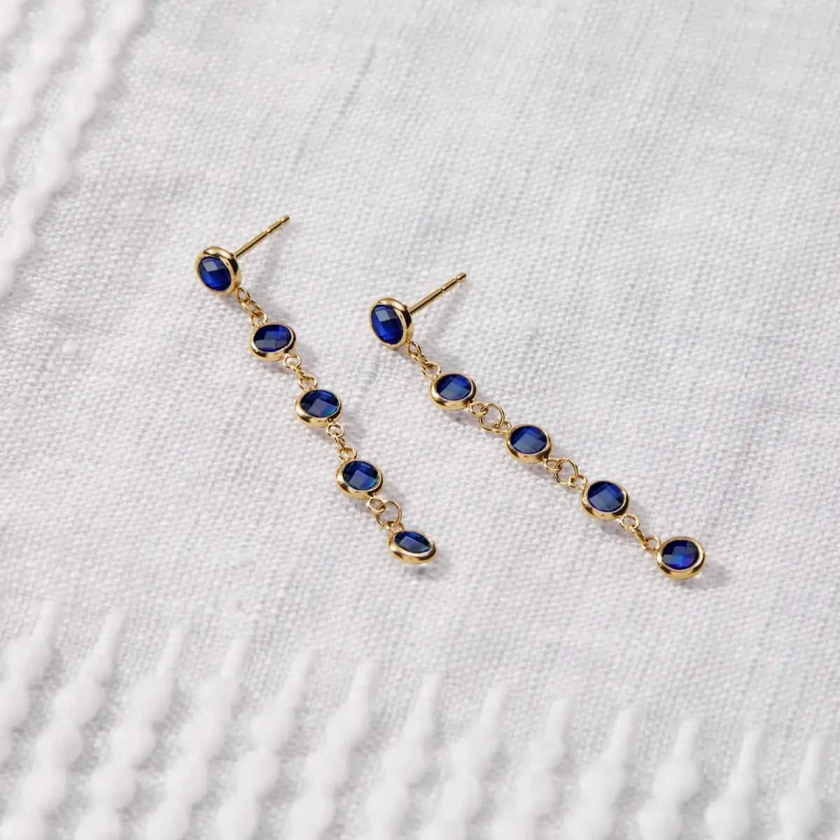 Pair of Newport Sapphire Earrings in 14k gold, each featuring nine bezel-set blue sapphires, displayed on textured fabric.