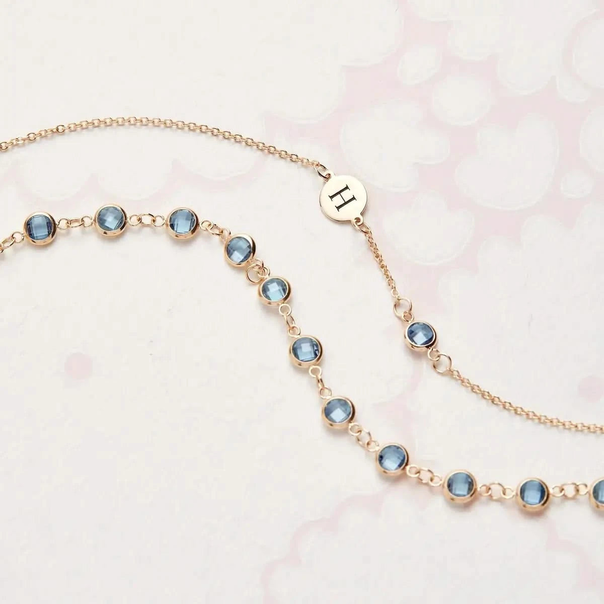 Flatlay of two 14k gold Nantucket Blue Topaz necklaces, one with a personalized initial charm and bezel-set stones.