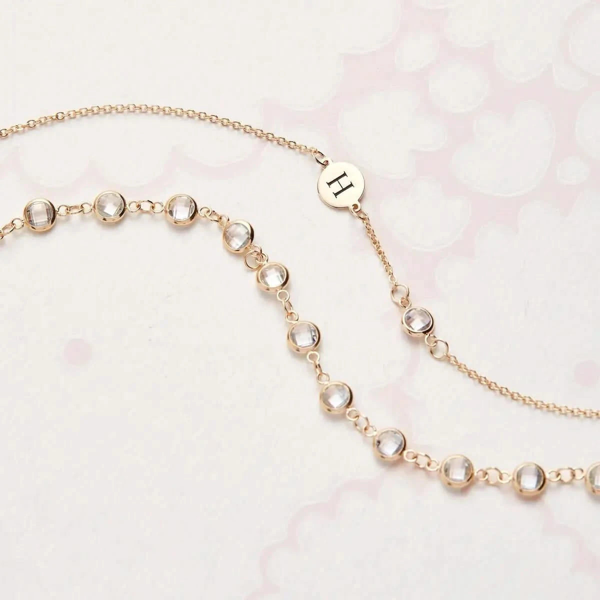 Detail view of the Newport White Topaz Necklace featuring bezel set round gemstones and an engraved H letter disc on a pink patterned background.