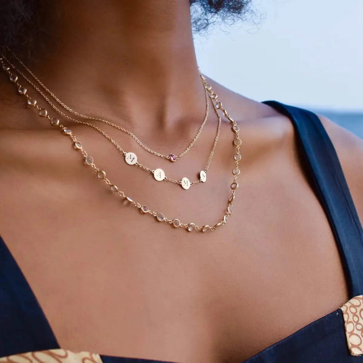 Woman wearing a layered necklace look featuring the Newport White Topaz Necklace in 14k gold styled with an engraved MAMA necklace and a single birthstone necklace.