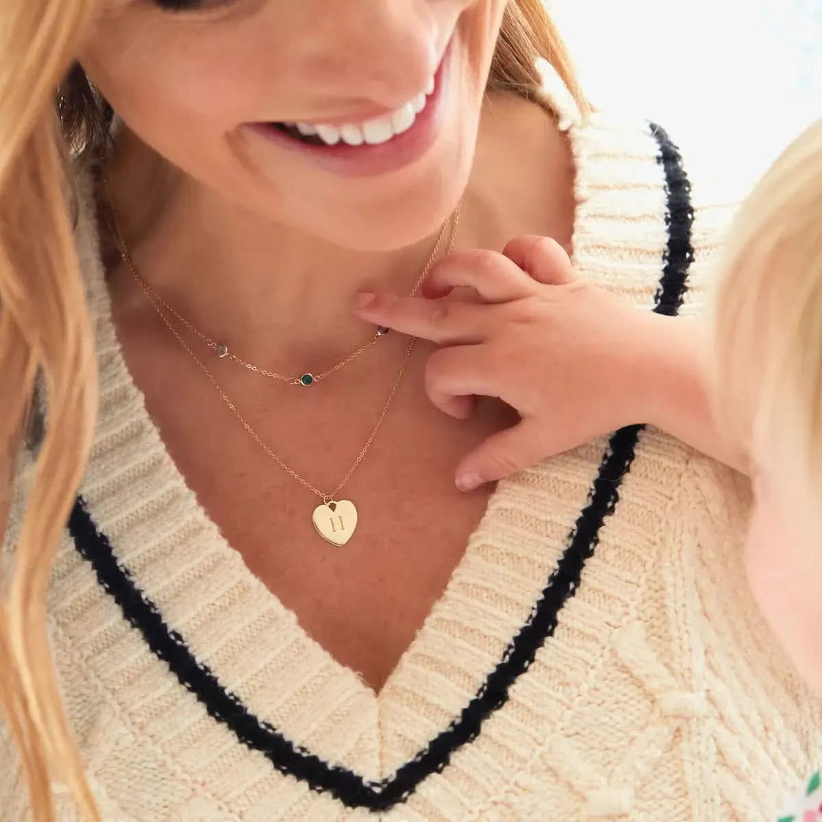 Child touches a gold heart pendant engraved with "H", worn on a classic chain necklace layered with a birthstone strand.