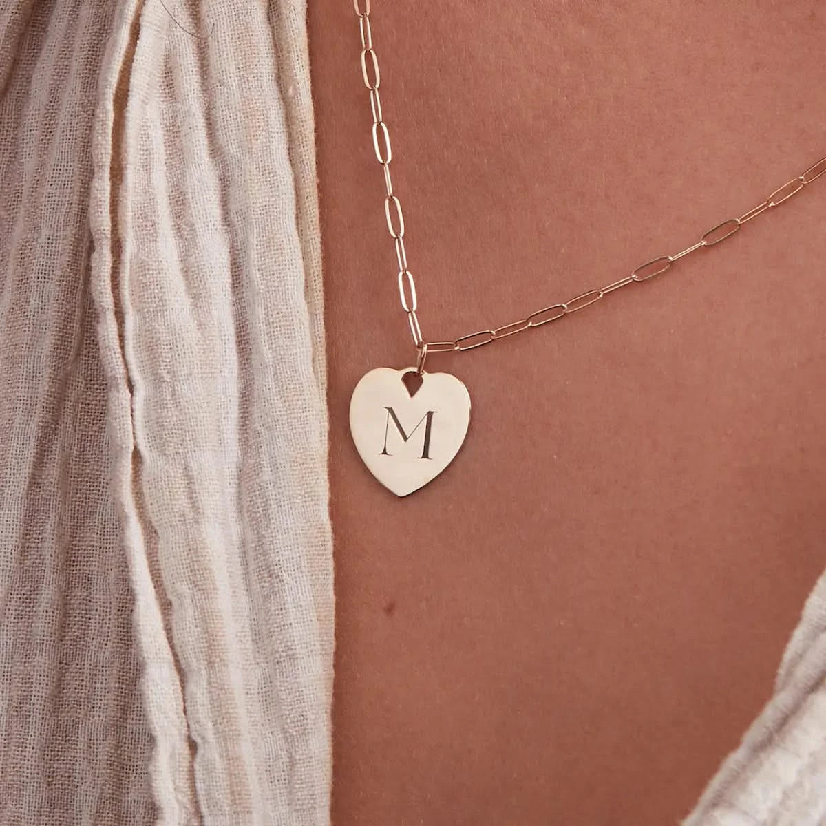 Close-up of 14k gold heart pendant engraved with the letter M on an Adelaide Mini chain, styled with a textured cream top.