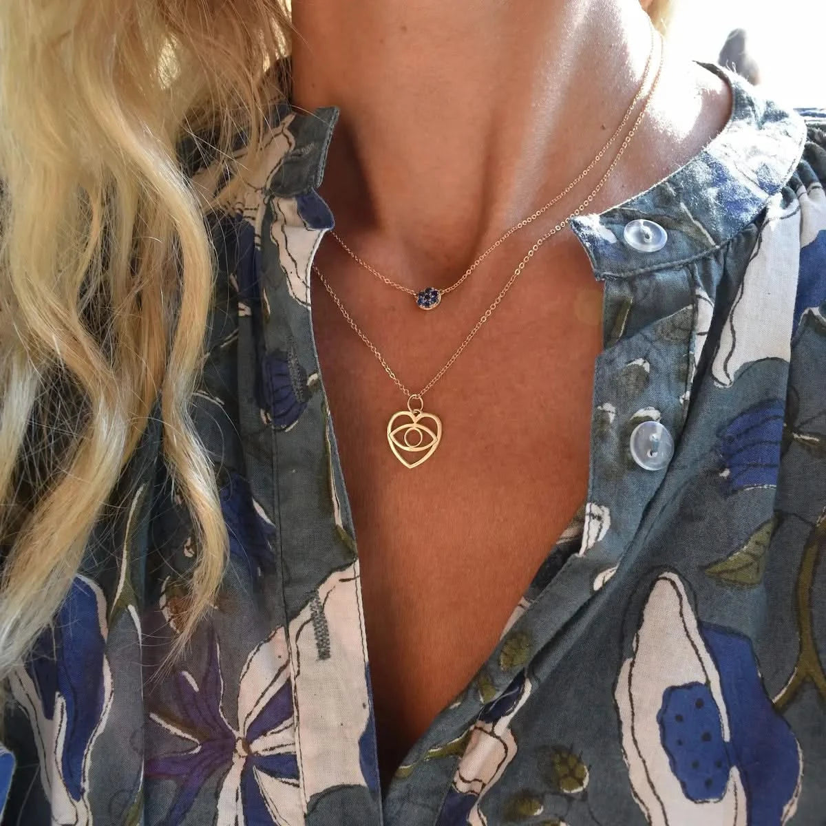 Small 14k gold heart pendant with cut-out evil eye symbol worn on a layered chain, styled with a floral blouse to show its lightweight, everyday wear appeal.