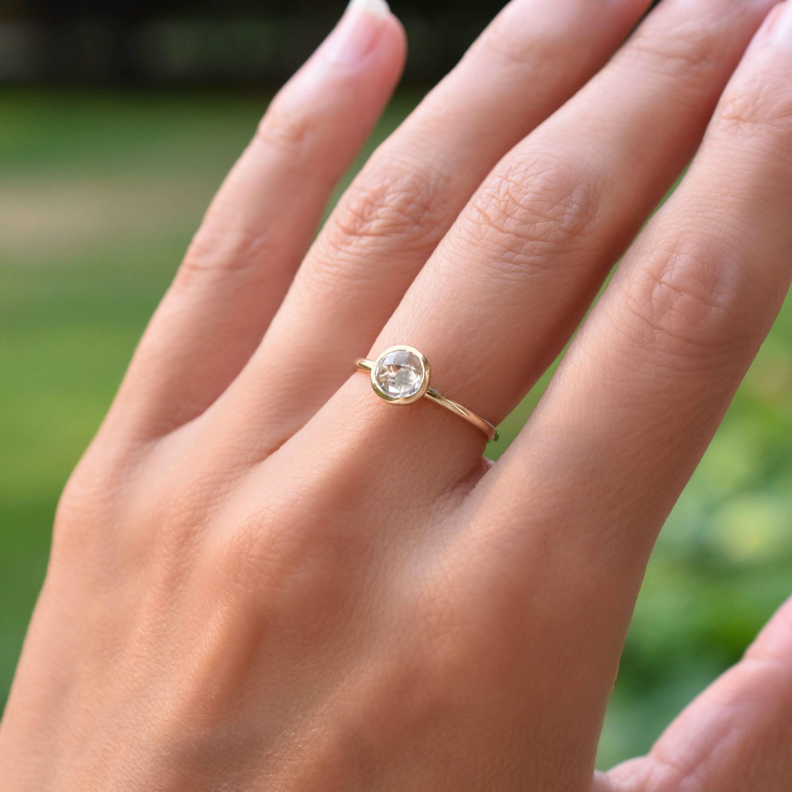 Hand wearing a 1.6 mm wide 14k yellow gold Grand ring featuring one 6 mm briolette cut bezel set white topaz