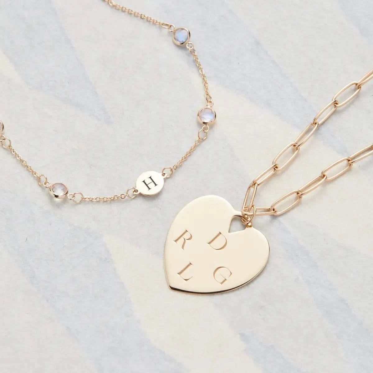 Flatlay of engraved Large Flat Heart Pendant in 14k gold paired with a moonstone necklace featuring an engraved H disc.