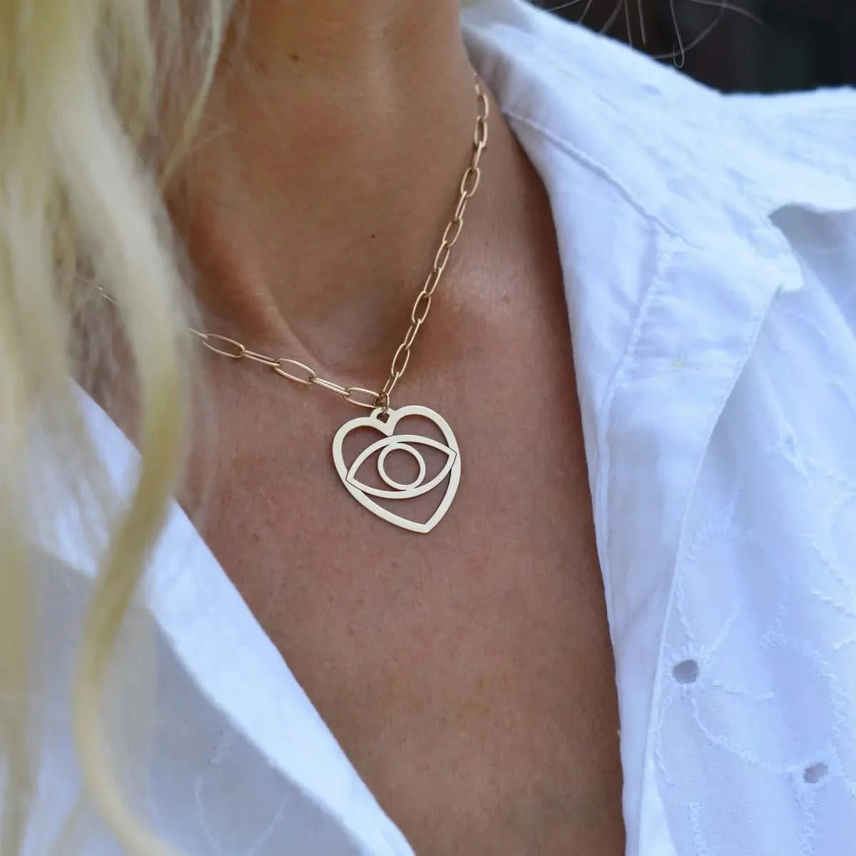 Yellow gold large Evil Eye Heart Pendant worn outdoors with a white eyelet blouse, shown in natural light.
