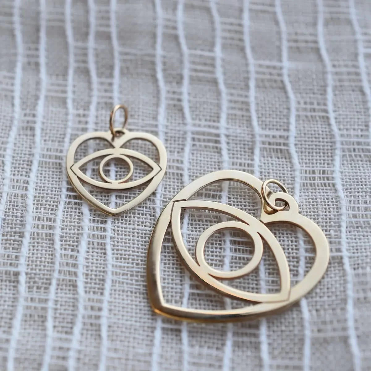 Small and large yellow gold Evil Eye Heart Pendants placed side by side on textured fabric.