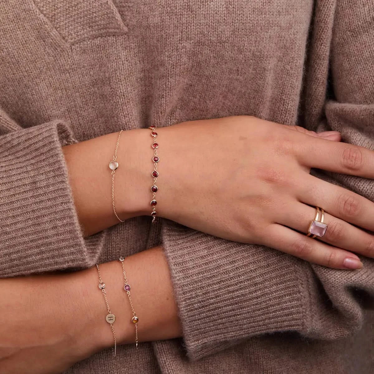 Woman wearing layered 14k gold birthstone bracelets and stacking rings, styled with a cozy knit sweater.
