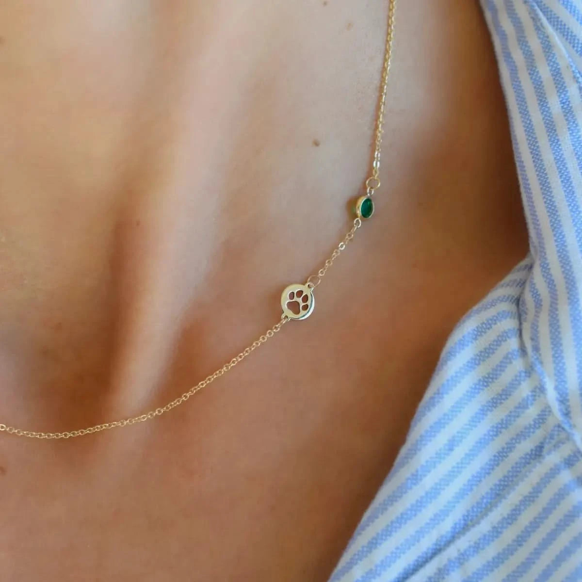 The gold paw cutout charm and bezel set emerald birthstone are worn by a woman, positioned close to the neck.
