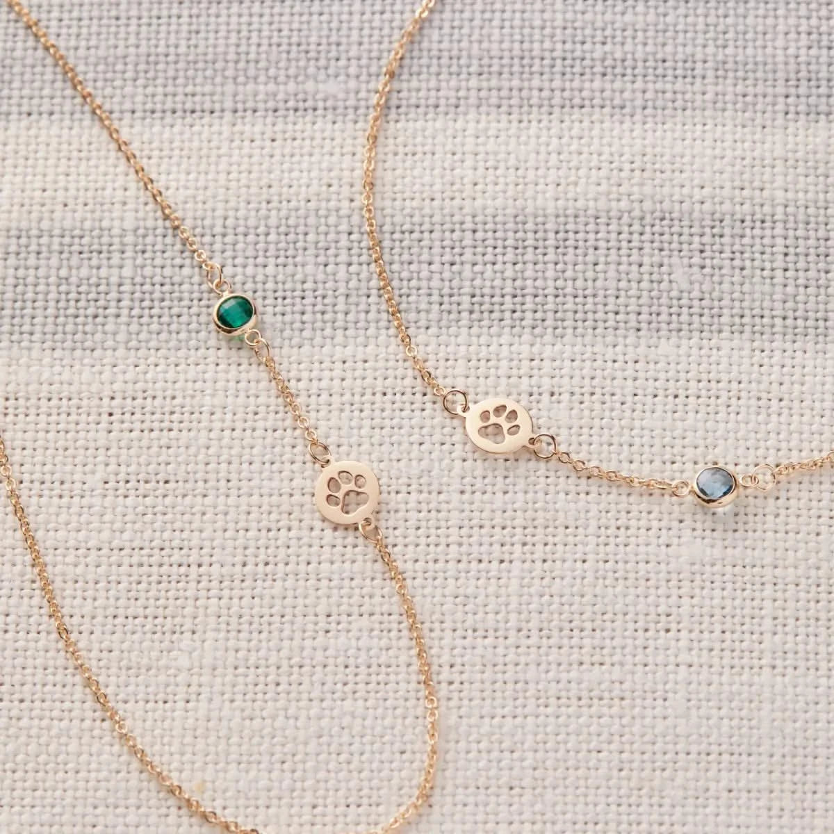 An asymmetrical Petite Birthstone necklace and matching Paw & Petite Birthstone bracelet in 14k gold are shown laid flat.