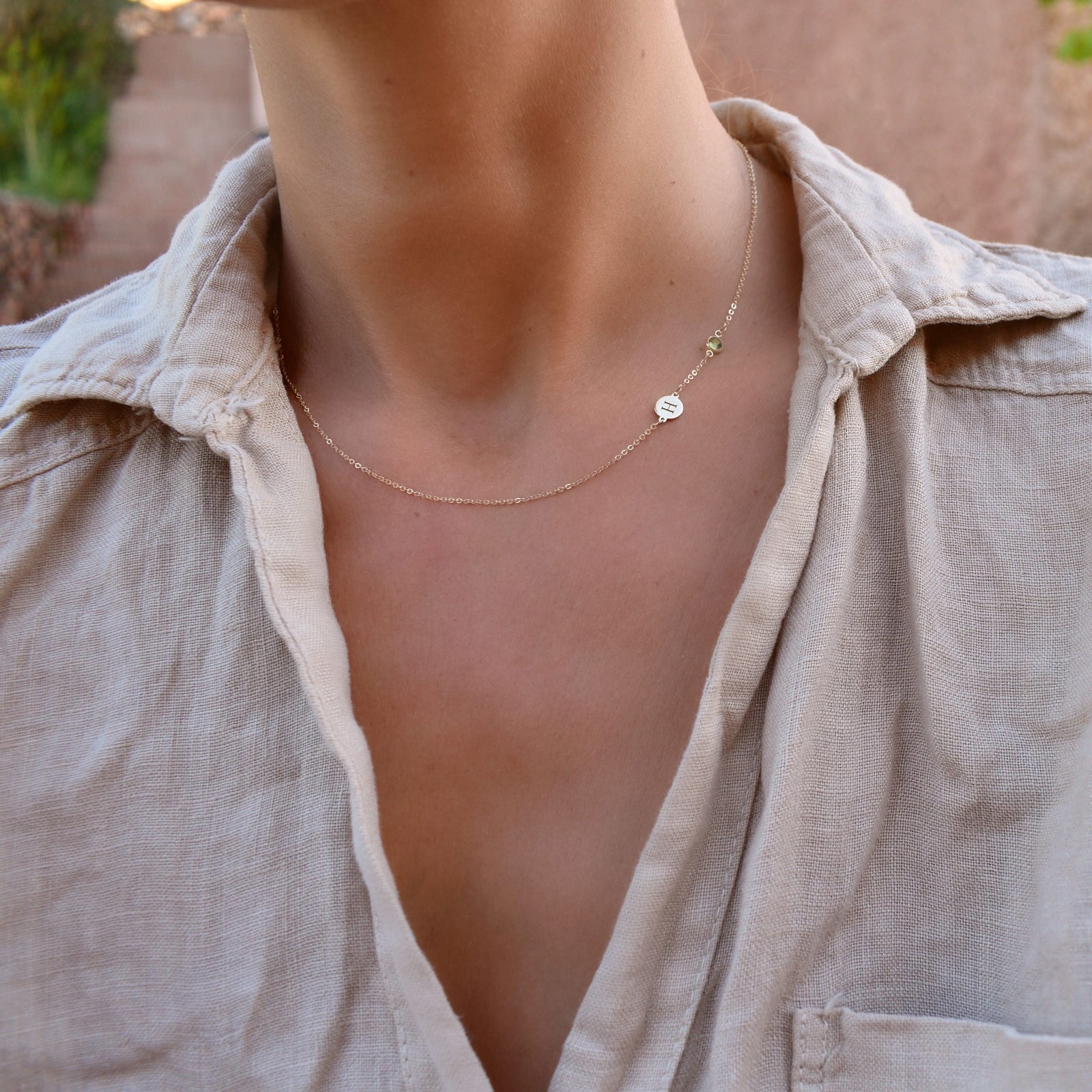 Woman wearing a 14k yellow gold Classic necklace featuring one peridot and one 1/4” flat disc engraved with the letter H