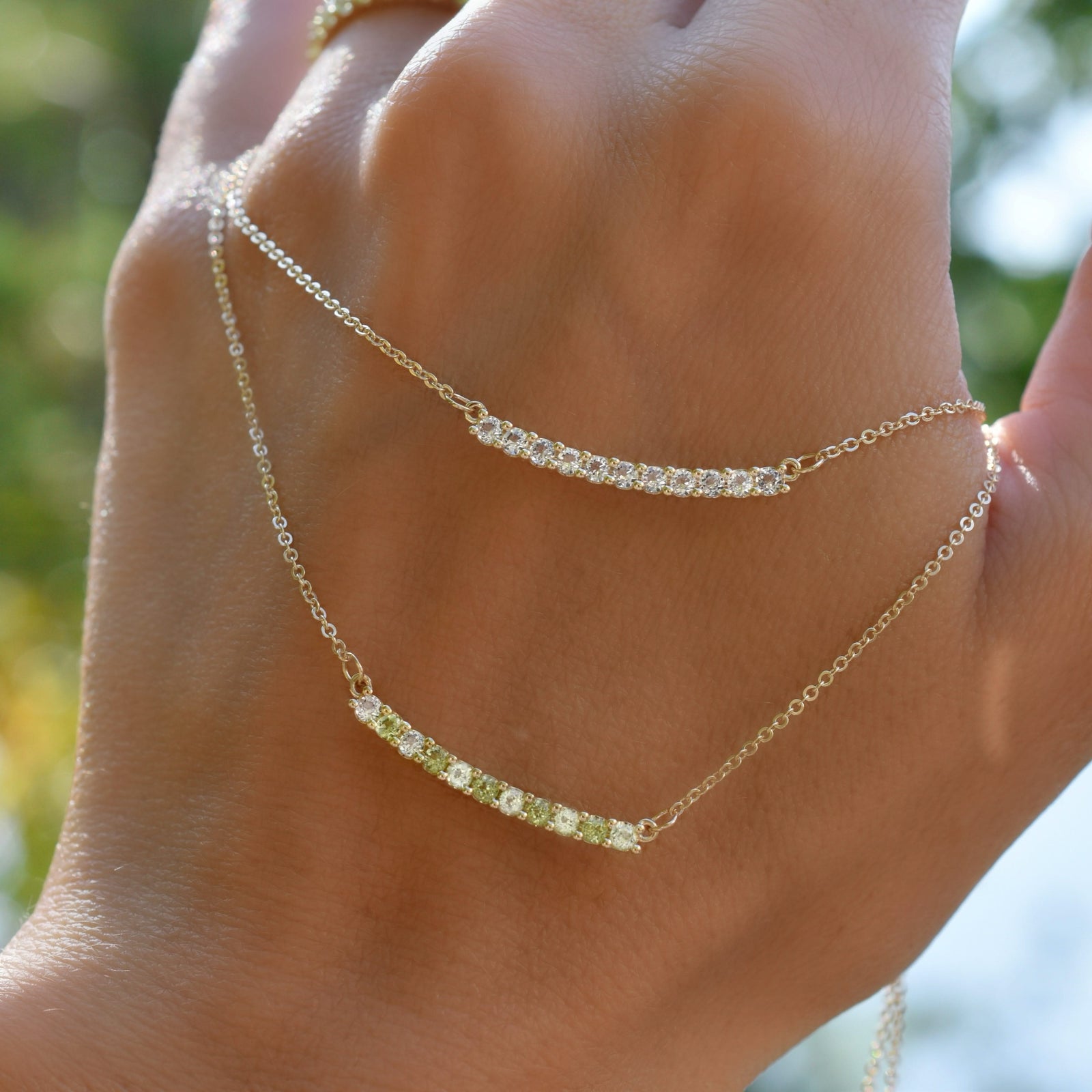 Hand holding two Rosecliff bar necklaces, one with eleven alternating 2 mm peridots and diamonds prong set in 14k gold