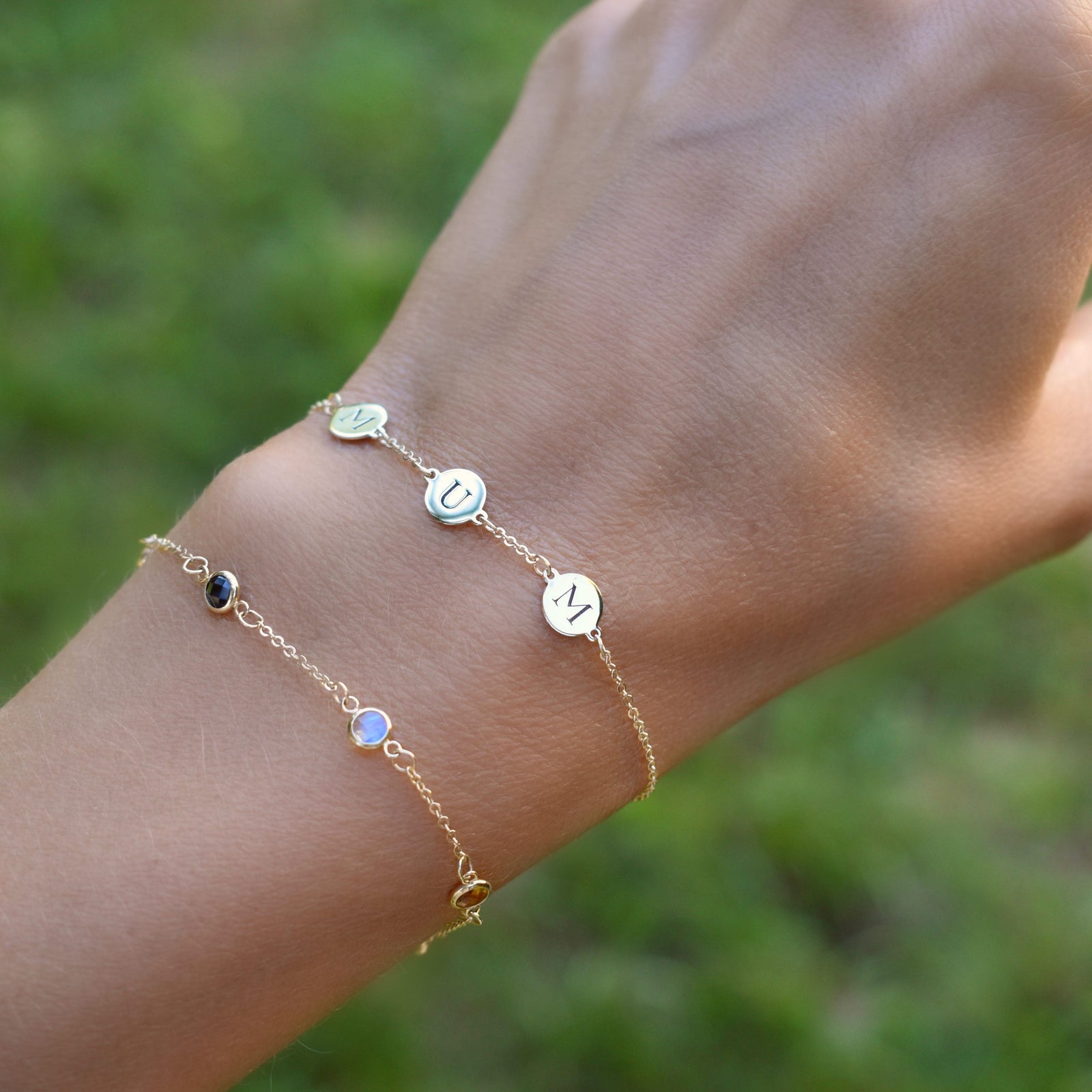 Hand wearing two bracelets including a 14k gold bracelet featuring three 1/4” flat engraved letter discs, spelling MUM