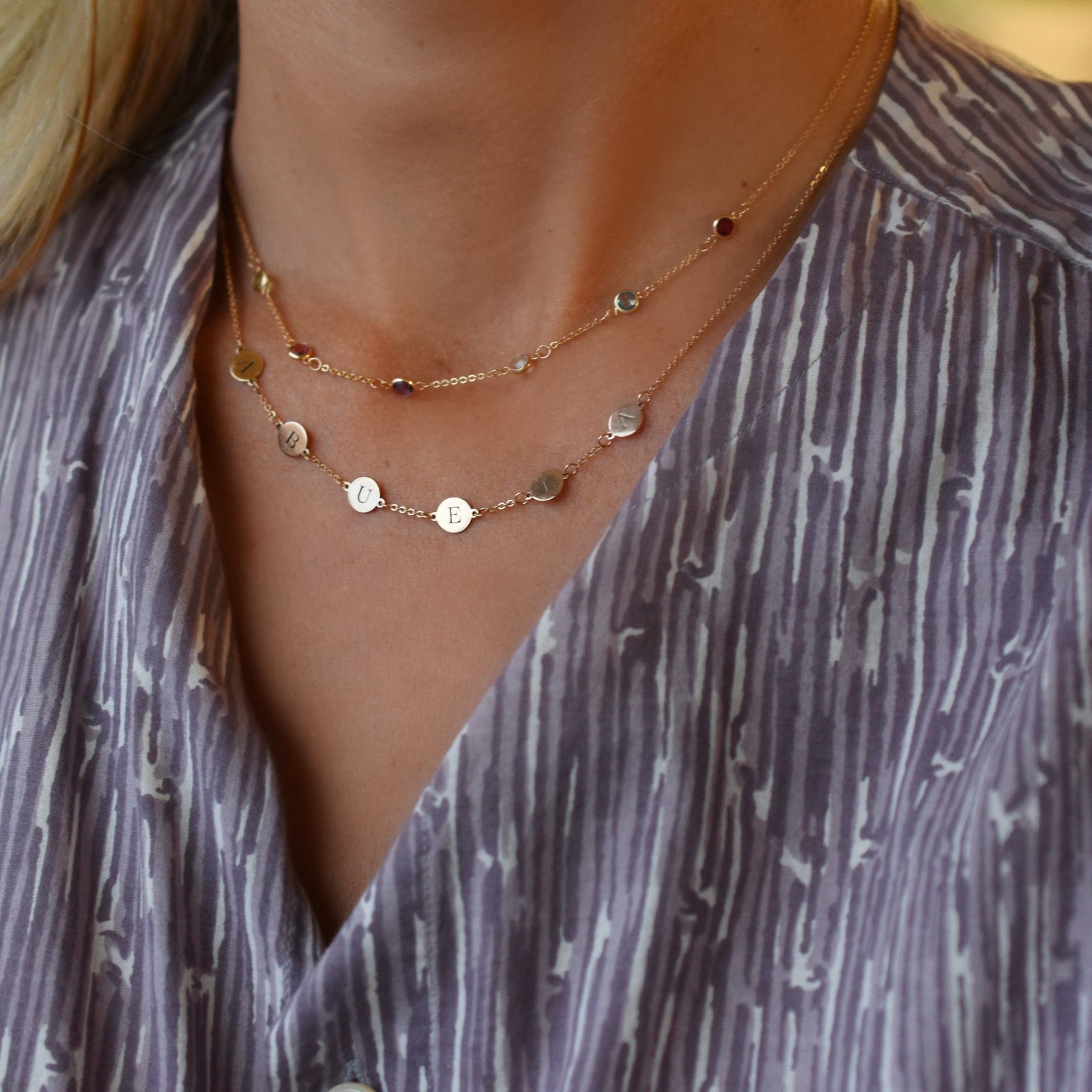 Woman wearing two necklaces including a 14k gold necklace featuring six 1/4” flat engraved letter discs, spelling Abuela