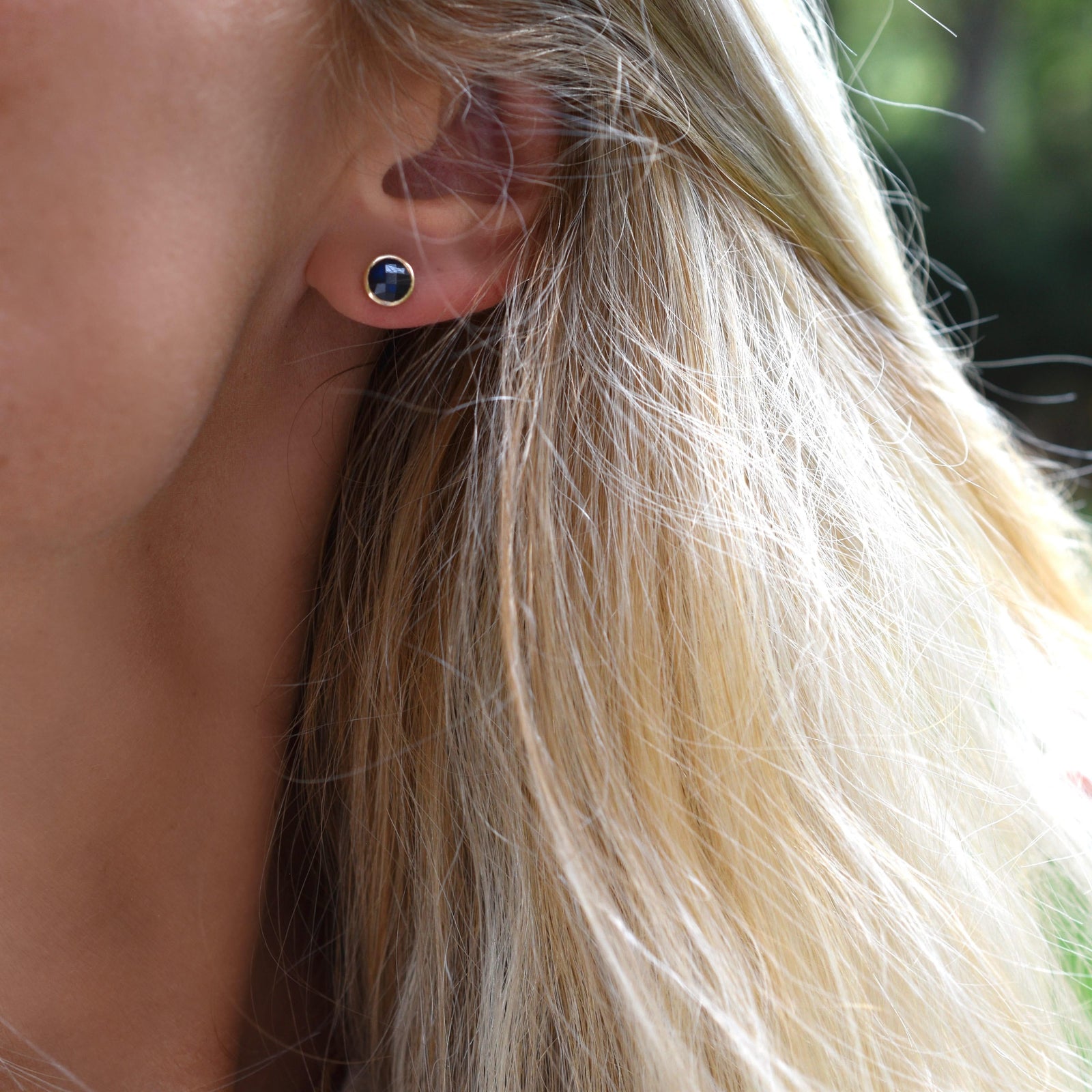 Woman wearing a 14k yellow gold Grand stud earring featuring one 6 mm briolette cut bezel set sapphire