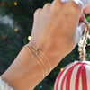 Woman's hand raised to hang ornament on a Christmas tree. On her wrist she is wearing 3 HAVERHILL Birthstone bracelets. Each bracelet is 14k yellow gold and has a dainty chain with a single birthstone in the center (emerald, blue topaz & aquamarine
