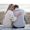 Couple sitting on a blanket on the beach in sweaters. Woman has her arm around the man's back and she is whispering in his ear.