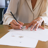 A woman sketches a jewelry design on paper while sitting at a desk.