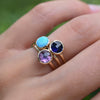 Finger wearing three stacked Grand rings featuring Amethyst, Turquoise, and Sapphire, bezel set in 14k yellow gold.
