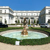 A fountain surrounded by manicured foliage in front of Rosecliff. People walk by in the background.