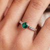 Woman's finger adorned with a 14k gold Rosecliff Grand Ring with a 6 mm round, briolette-cut Emerald and eight 2 mm round-cut Diamonds.