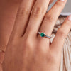 A woman's hand adorned with the Rosecliff Grand Emerald Ring in 14k gold, showcasing a round, briolette-cut, bezel-set emerald flanked by accent stones perfect for engagement.