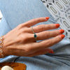 Woman's hand wearing Haverhill 14k gold rings, including a 14k gold Rosecliff Grand Ring with a 6 mm briolette-cut Emerald and eight smaller 2 mm round-cut Emerald.