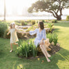 A young daughter runs playfully around a rock that her smiling mother is sitting on while they hold each other's hand.