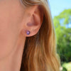 Woman wearing a pair of Grand Birthstone Stud Earrings in 14k Gold featuring one 6 mm briolette cut, bezel set Amethyst. 