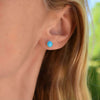 Woman showcasing a pair of 14k yellow gold Grand stud earrings each featuring one 6 mm briolette cut bezel set turquoise.