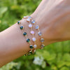 Woman wearing 3 layered Newport Grand bracelets in 14k yellow gold featuring aquamarine, pink opal, and emerald gemstones.