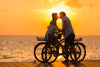 A couple standing with bicycles on a dock exchange a kiss in front of the setting sun.