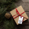 A brown gift box tied with a red ribbon with a card saying, 'Open Me.' Austrian Pine boughs and a pinecone lay to the left.