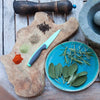 Wood live edge cutting board with knife and herbs, blue plate with herbs, and stone mortar and pestle set on a wooden table.