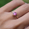 14k yellow gold Grand ring featuring a 6mm briolette cut, bezel set pink sapphire gemstone on a 1.6mm band, worn on a finger.